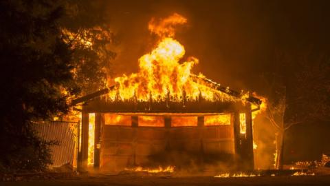 A garage door, buckled and collapsed off its hinges, lies resting against the frame of a garage engulfed in flames in this dark shot of a burning home, the air black with smoke and nearby trees also catching fire