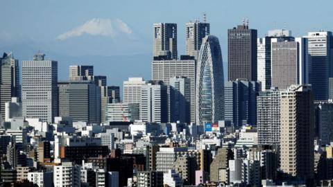 A snow-topped Mount Fuji behind the Shinjuku skyline in Tokyo, crowded with skyscrapers on a sunny day.