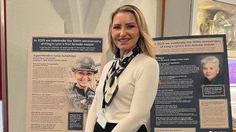 Supt Sonia Humphreys stands in front of a display board at the Stories of Lynn Museum. She has long blonde hair and wears a white top and black and white scarf. 