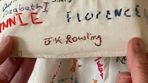 A pair of hands are holding a piece of fabric covered in embroided names. The one being held prominently to the camera is "JK Rowling"