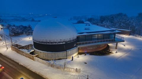 A domed planetarium building seen from above in the snow as darkness is falling