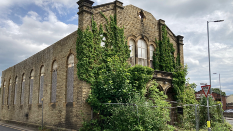 An old, run-down looking building with large circular windows on the side. There is greenery covering part of the exterior and a metal fence bordering some of the building. 