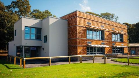 Brick and grey render building with 9 windows and a door. There are trees and grass.