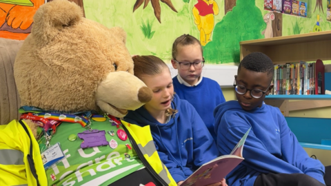 Three primary school children - two girls and a boy - are wearing blue uniform hoodies and sitting reading a picture book with a beige-furred, human-sized cuddly toy bear in a school library. Beside them is a bookshelf and on the wall behind them is a mural of Winnie the Pooh in a green-grassed forest with trees.