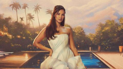 Maya Jama in a promo shot for the new series of Love Island All Stars. Maya wears a white dress and poses with her hand on her hip in front of a pool and palm trees. The shot has been edited to look like a painting, with soft summer lights and orange hues. 