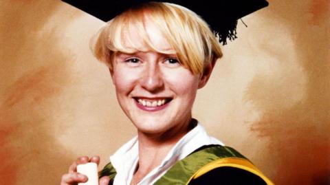 A graduation picture of Melanie Hall, wearing a white collared shirt under green and yellow robes with a mortar board. She has short blonde hair and blue eyes, and is smiling at the camera while holding a scroll. The background is various hues of orange.