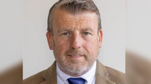 A head and shoulders profile picture of councillor David Maclean, who has short grey hair and beard, is wearing a brown suit, white shirt and blue tie, against a white background