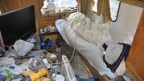 Interior of caravan strewn with old bedding, dirty cooking utensils and electrical equipment