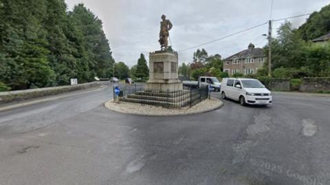 A van is seen going around a roundabout with a statue in the middle.