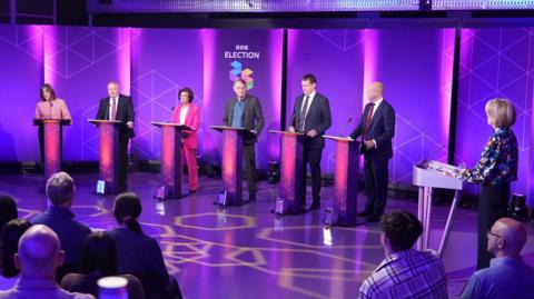 The six party leaders and host Bethan Rhys Roberts standing at lecterns on the purple studio set with BBC election branding behind them.