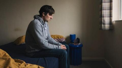 A wearing a grey hood and jeans is sitting on a bed. He has a sad look on his face and he is looking down at the floor, with his hands clenched.