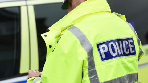 A male police officer wearing a florescent police jacket while standing in front of a police car 