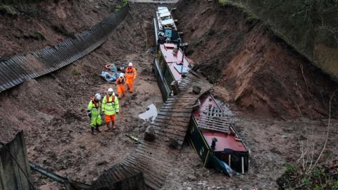 Two boats at the bottom of a hole in a canal with no water in. one is sunk partially into the earth and has a wooden fence lying over the top of it. there are four people standing in the hole next to them in fluorescent yellow and orange clothing with white hard hats
