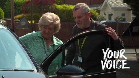 A man with grey hair who is wearing a yellow lanyard opens the door for an elderly woman in a green top. 