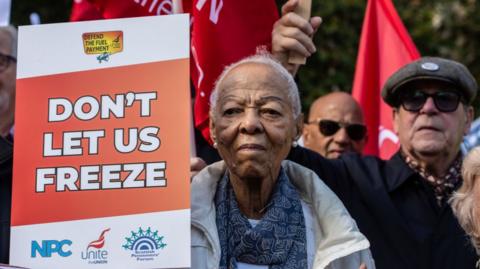 Pensioners attend the protest at Parliament on 7 October over the decision to cut the winter fuel payment to some. A woman is holding a placard and a man can be seen in the background with a flag
