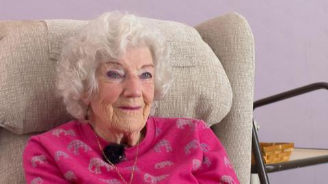 Nora Hoskin, sat on a cream armchair, she is weaing a pink jumper with a gold necklace, she is facing the camera directly. The wall behind her is purple. 