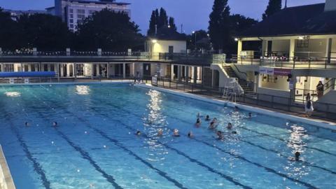 A large swimming pool with surrounding buildings, which are lit because it is in the evening. A group of swimmers gather in the pool.