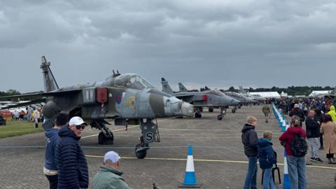 A number of fighter jets lined up in a taped off area on an airfield. People have gathered around to look at the aircraft.