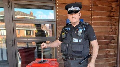 A police officer wearing a black t-shirt and black vest that says "police" on it, in silver writing on a blue background. There is a walkie talkie on his chest, a body cam, and a baton strapped to his waistband. He is holding a small knife over the entrance of a red knife surrender bin.