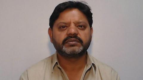 A mugshot of Kasir Bashir shows him staring directly at the camera with no expression on his face. He has black hair, a black mustache and a greying beard, and is wearing a beige shirt.