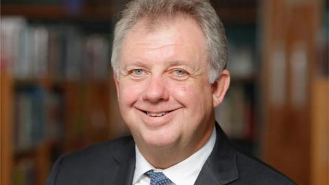 Sidwick has short grey hair and wears a suit in an official wood-panelled setting.