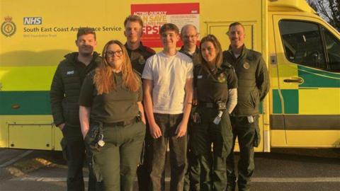 Six South East Coast Ambulance crew members standing around a teenage boy who is wearing a white shirt and grey jeans. They are standing in front of an ambulance.