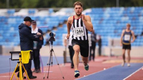 Evan Campbell runs down the blue long jump track in mid-air, wearing black and white vertical striped vest and thigh-length shorts