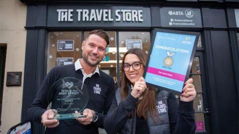 A man and a woman standing in front of a shop called The Travel Store. The man is holding an award and the woman is holding a framed certificate.