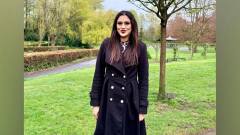 Councillor Simran Cheema stood smiling in a park. She has long shoulder length brown hair, wears red lipstick and a double breasted black coat.