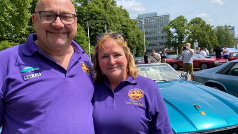 A man and a woman both wearing purple tops stand in front of a turquoise car