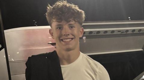 A teenage boy with curly blond hair smiles as he stands in front of a parked silver van at night. He is wearing a white T-shirt and has a black jumper chucked over one of his shoulders.