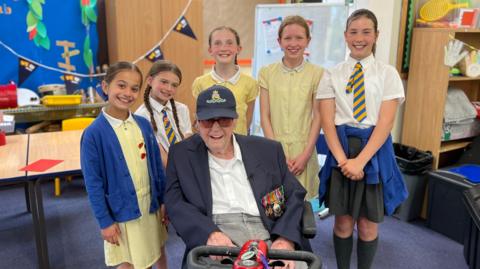 A smiling Mr Jones is sitting on a mobility scooter, wearing a navy jacket, grey trousers, glasses, and a blue cap. Around him are five smiling students from the school. They are all in a classroom setting.