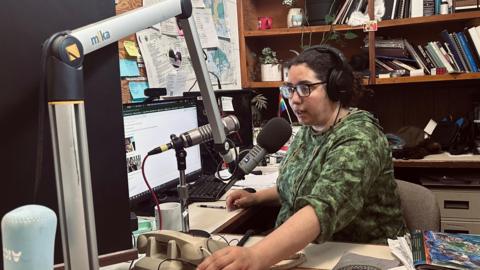 Desire Hagan, wearing a green sweatshirt, glasses and earphones, speaks into a microphone in a radio studio.
