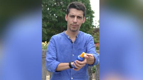 A man in a blue linen shirt applies sunscreen. He has the blue and yellow bottle in his hand.