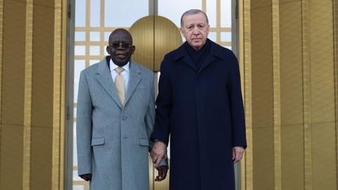 Bola Tinubu in a long grey coat is standing next to his Turkish counterpart in a long black coat. Erdogan is holding Tinubu's wrist. They are standing on a turquoise carpet and a Turkish flag can be seen on the right.