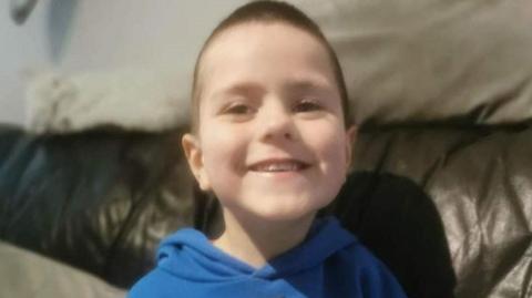 Young boy smiling into the camera. He is wearing a blue hoodie and sitting on a brown leather sofa.