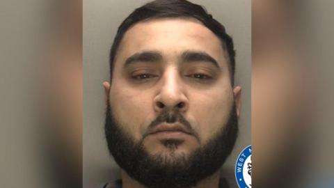 A police mugshot of a man with short black hair and a large black beard.