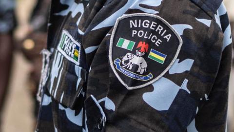 A close-up of a badge saying "Nigeria Police" on the sleeve of a camouflage police uniform. The police's emblem of an elephant with a red eagle above is also on the badge.