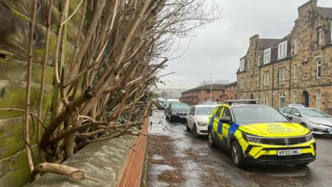 A wet residential street with a low wall and wooden fence on the left and the branches of a hedge in-between. A police car is parked in the foreground.