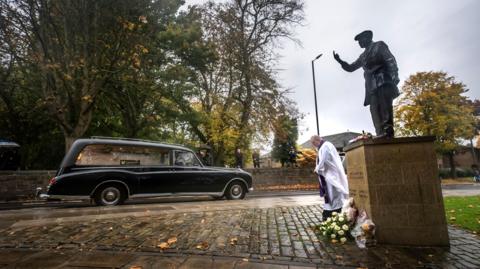 A hearse passing a statue with mourners lining the route