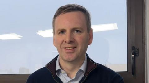 Colin Broomfield, Director of Markets at the Utility Regulator, smiling to the camera standing at a window. He is wearing a light blue shirt and navy zip top up jumper. His hair is short and grey.