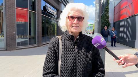 A woman with white hair and sunglasses and a black jacket being interviewed by a radio reporter in a shopping centre