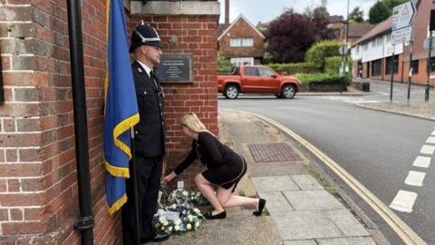 A woman in black kneels down to lay some flowers outside a building. 