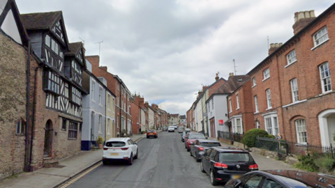 A street with rows of houses on both sides, one of them with timber frames and cars parked on both sides