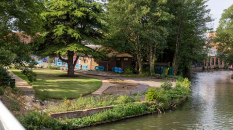 Newbury Peace Garden sits alongside a canal. It has grass trees and pathways - a number of blue barriers can be seen next to a building in the background.