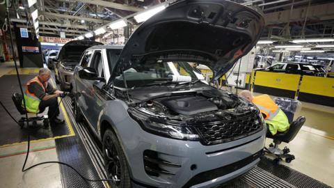 A car manufacturing assembly line featuring a grey SUV in the foreground with its hood open. Two workers are seated on either side of the vehicle - one is working on the rear wheel on the left side, the other is by the front wheel on the right. The background includes additional vehicles in various stages of production, along with industrial machinery and equipment. The factory is brightly lit with overhead lights.