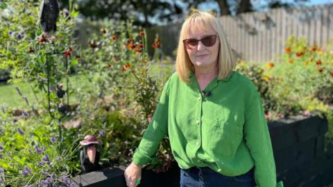 A woman wearing a green buttoned shirt. She is wearing sunglasses and in the background is a colourful garden.