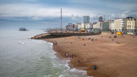 Visitors on a pebbled beach and grey skies. There is the sea to the left and a beach to the right. 