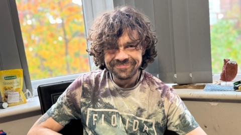 A man sat in a window with curly brown hair. 