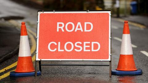 A red road closed sign with white lettering. It is in the middle of a road with a orange and white traffic cone on either side